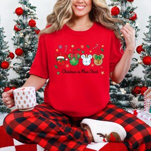 May include: A red t-shirt with a festive Christmas design, including Mickey Mouse-shaped ornaments and the words "Christmas on Main Street." The shirt is worn with red and black checked pyjama bottoms.