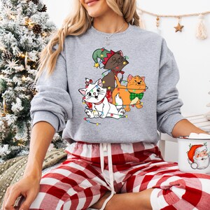 May include: A gray sweatshirt featuring a graphic of three cartoon cats decorated with Christmas lights and festive accessories. The cats are a white cat with a red bow, a brown cat with a green hat, and an orange cat with a green bow tie. The person is wearing red and white plaid pajama pants.
