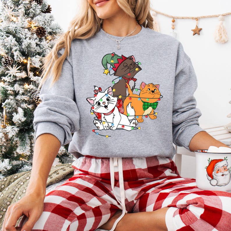 May include: A gray sweatshirt featuring a graphic of three cartoon cats decorated with Christmas lights and festive accessories. The cats are a white cat with a red bow, a brown cat with a green hat, and an orange cat with a green bow tie. The person is wearing red and white plaid pajama pants.