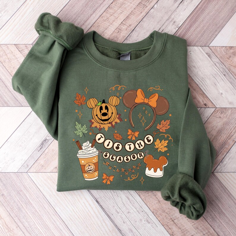 May include: Green sweatshirt with a festive Halloween design. The design includes a pumpkin Mickey Mouse, fall leaves, a coffee cup, and the phrase "'Tis the Season". The sweatshirt has long sleeves and a crew neck.