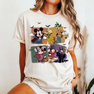 K&ouml;nnte beinhalten: Ein cremefarbenes T-Shirt mit einer Vintage-Grafik von Mickey Mouse, Minnie Mouse, Donald Duck, Goofy und Pluto in Halloween-Kost&uuml;men. Die Grafik ist von Flederm&auml;usen und K&uuml;rbissen umgeben.