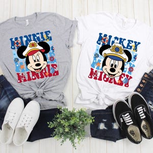 May include: Two gray and white t-shirts with a nautical theme featuring Mickey and Minnie Mouse wearing captain's hats. The shirts have the text "Mickey Mouse" and "Minnie Mouse" on them. The shirts are displayed on a white wooden surface with blue jeans and white sneakers.