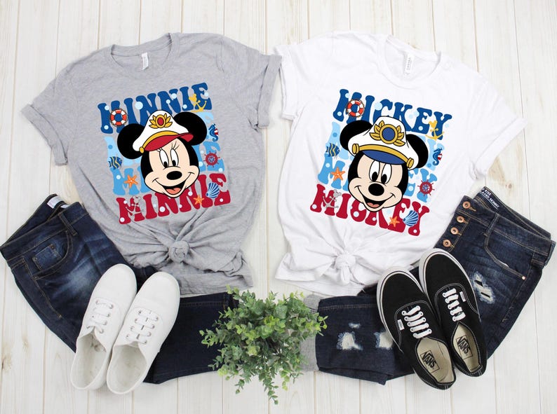 May include: Two gray and white t-shirts with a nautical theme featuring Mickey and Minnie Mouse wearing captain's hats. The shirts have the text "Mickey Mouse" and "Minnie Mouse" on them. The shirts are displayed on a white wooden surface with blue jeans and white sneakers.
