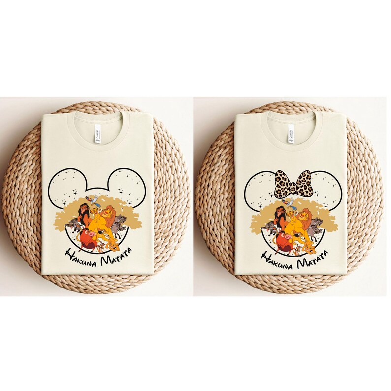 Puede incluir: Dos camisetas de color crema con un gr&aacute;fico de los personajes de El Rey Le&oacute;n dentro de una silueta de cabeza de Mickey Mouse. Una camiseta tiene un lazo con estampado de leopardo. Ambas camisetas tienen el texto "Hakuna Matata" impreso debajo del gr&aacute;fico.