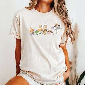 Op de afbeelding: Een beige T-shirt met een afbeelding van Toy Story-personages die verkeerskegels dragen. De personages zijn Woody, Buzz Lightyear, Mr. Potato Head, Rex en Hamm.