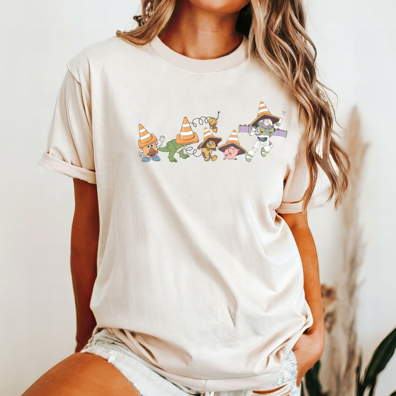 Op de afbeelding: Een beige T-shirt met een afbeelding van Toy Story-personages die verkeerskegels dragen. De personages zijn Woody, Buzz Lightyear, Mr. Potato Head, Rex en Hamm.