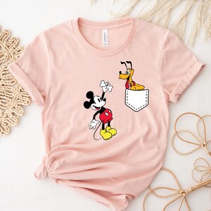 Puede incluir: Una camiseta rosa con un bolsillo blanco que presenta a Mickey Mouse sosteniendo un hueso y a Pluto asom&aacute;ndose desde el bolsillo.