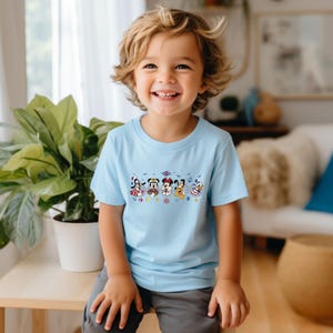 May include: A light blue t-shirt featuring a colorful graphic of Disney characters. The graphic includes Mickey Mouse, Minnie Mouse, Donald Duck, and other characters. The shirt is worn by a young child.
