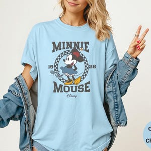 May include: Light blue t-shirt featuring a vintage-style graphic of Minnie Mouse. The design includes the text "MINNIE MOUSE" and "1928". The shirt is a comfort colors chambray.