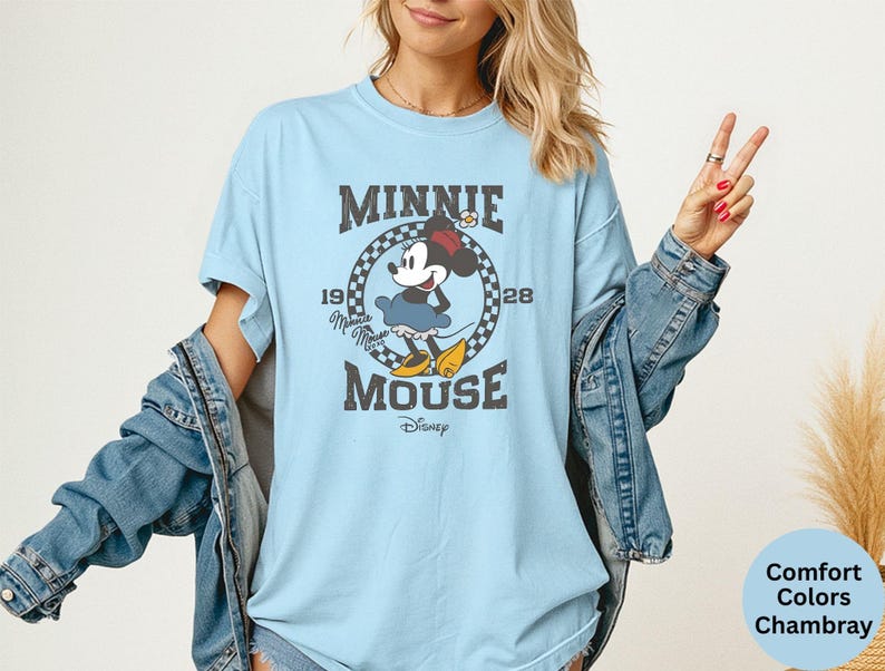 May include: Light blue t-shirt featuring a vintage-style graphic of Minnie Mouse. The design includes the text "MINNIE MOUSE" and "1928". The shirt is a comfort colors chambray.