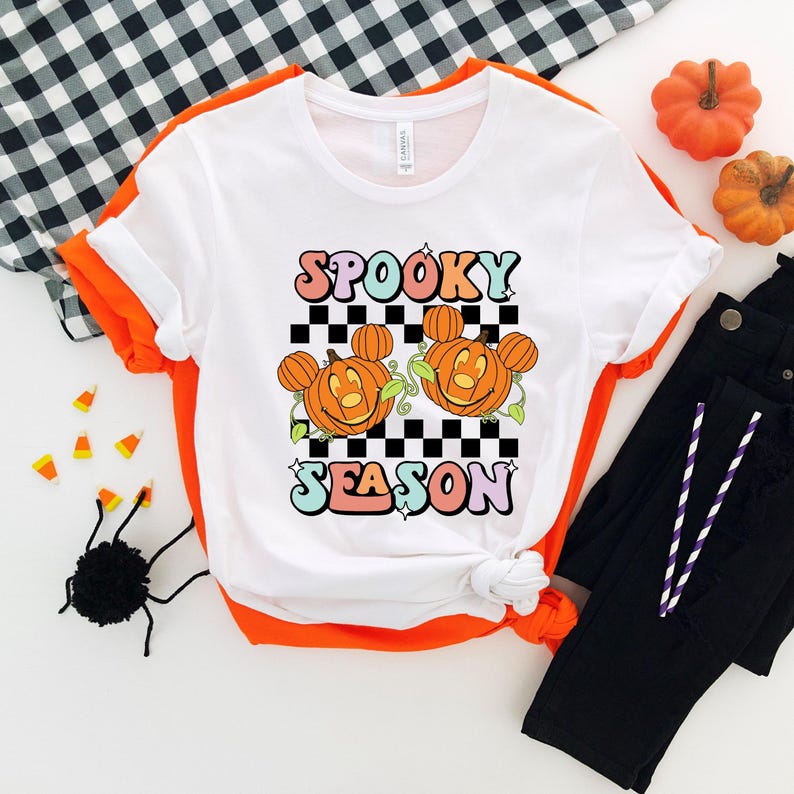 May include: White t-shirt with orange trim featuring the words "Spooky Season" in colorful letters. The design includes two smiling jack-o'-lanterns with Mickey Mouse ears on a black and white checkered background. Halloween-themed apparel.