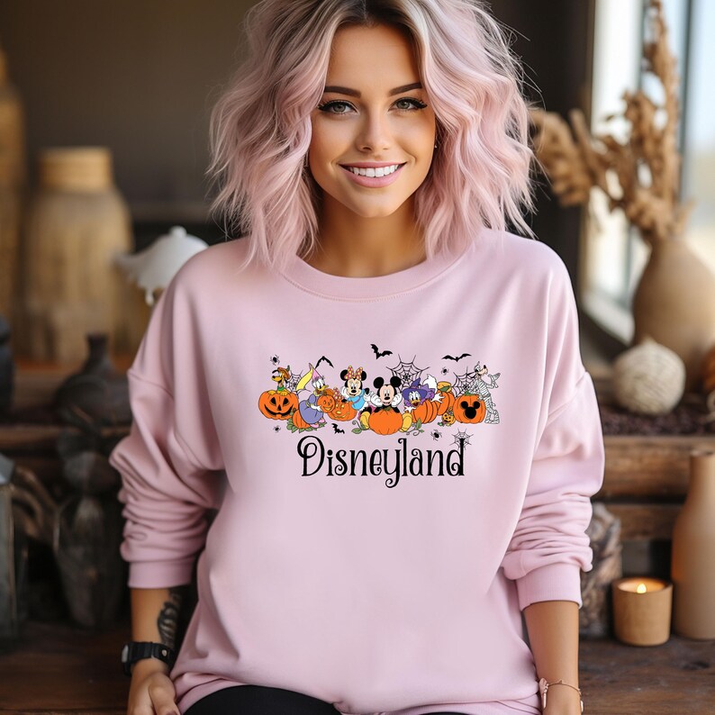 K&ouml;nnte beinhalten: Hellrosa Disneyland-Sweatshirt mit Halloween-Motiv. Die Grafik zeigt Zeichentrickfiguren, K&uuml;rbisse, Flederm&auml;use und das Wort "Disneyland". Das Sweatshirt hat lange &Auml;rmel.