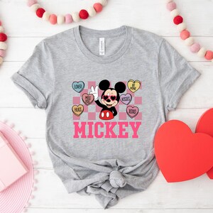 May include: A light gray t-shirt featuring a graphic of Mickey Mouse wearing sunglasses and giving a peace sign. The design includes heart-shaped candies with phrases like "Kisses" and "Be Mine," and the name "Mickey" in pink.
