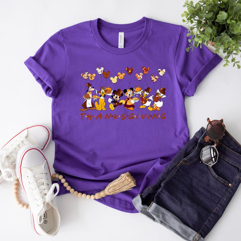 Pu&ograve; includere: Una maglietta viola con un design a tema Ringraziamento con personaggi Disney. Topolino, Paperino e Pippo sono raffigurati con palloncini. Il testo "THANKS GIVING" &egrave; stampato in arancione. La maglietta &egrave; abbinata a pantaloncini di jeans e scarpe da ginnastica bianche.