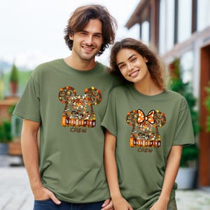 Puede incluir: Dos personas con camisetas verde oliva. Cada camiseta presenta un dise&ntilde;o de cabeza de Mickey Mouse con hojas de oto&ntilde;o y personajes, el texto "Thanksgiving CREW" y un lazo en una camiseta. Las camisetas son de corte holgado.