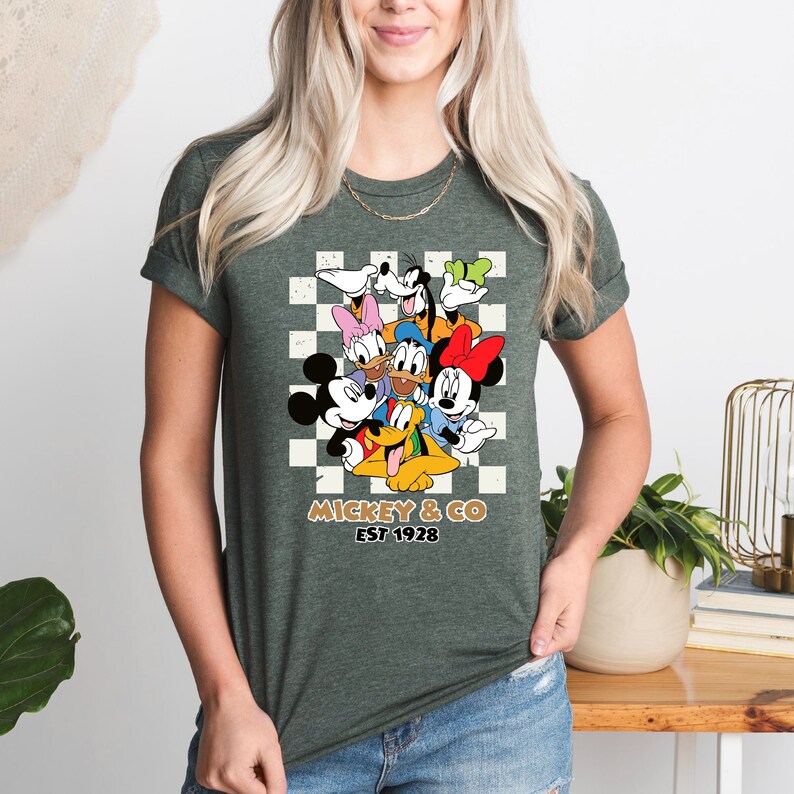 Puede incluir: Una camiseta verde con un fondo a cuadros con Mickey Mouse y sus amigos. El texto "Mickey & Co. Est 1928" est&aacute; impreso debajo de la imagen.