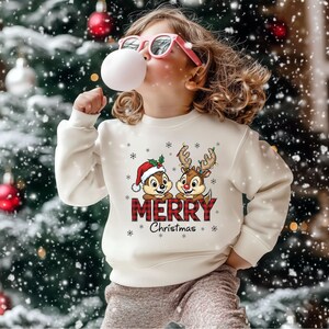 May include: A cream-coloured sweatshirt with a Christmas design featuring two cartoon chipmunks wearing a Santa hat and reindeer antlers. The word "MERRY" is in a red plaid pattern, with "Christmas" below. The child is wearing pink glasses and blowing a bubble.