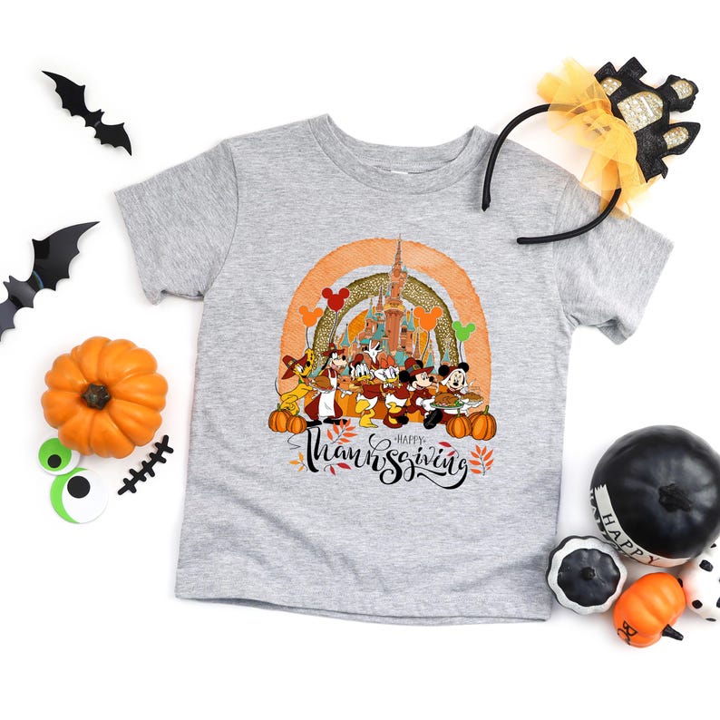 May include: A gray t-shirt featuring a colorful Thanksgiving design with Disney characters, a castle, and the words "Happy Thanksgiving." The shirt is surrounded by Halloween decorations, including bats, pumpkins, and a headband.