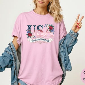 May include: A light pink t-shirt with the word "USA" in a patriotic design, featuring the American flag and floral accents. The text "250 Years of Freedom" is displayed on a banner below. The shirt is part of the Comfort Colors Blossom collection.