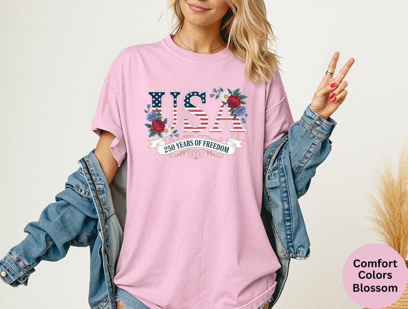 May include: A light pink t-shirt with the word "USA" in a patriotic design, featuring the American flag and floral accents. The text "250 Years of Freedom" is displayed on a banner below. The shirt is part of the Comfort Colors Blossom collection.