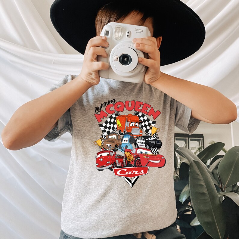 May include: A gray t-shirt with a graphic of Lightning McQueen and other Cars characters. The text "Lightning McQueen" and "Cars" are printed on the shirt.