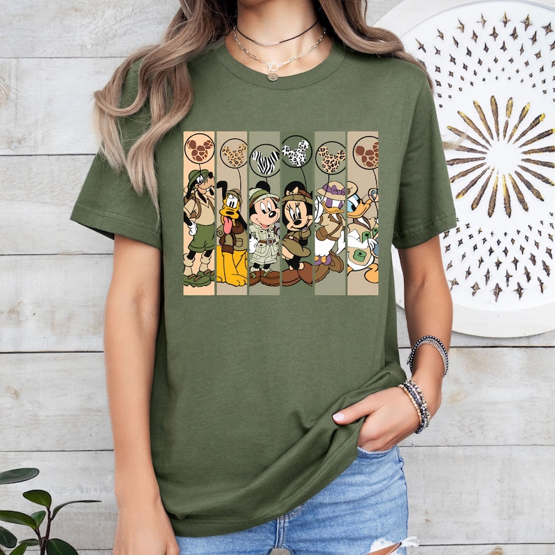 May include: A green t-shirt with a graphic print of Mickey Mouse, Minnie Mouse, Donald Duck, Goofy, and Pluto dressed in safari outfits. The characters are holding balloons with animal print designs.