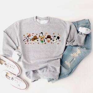 May include: A light gray sweatshirt featuring cartoon character faces with hats and fall leaves. The sweatshirt is paired with blue jeans and white sneakers.