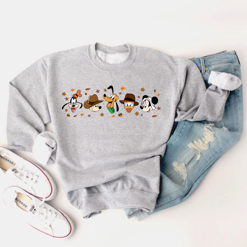 May include: A light gray sweatshirt featuring cartoon character faces with hats and fall leaves. The sweatshirt is paired with blue jeans and white sneakers.
