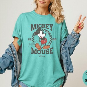 May include: Seafoam green t-shirt featuring a distressed graphic of Mickey Mouse. The design includes the text "Mickey Mouse" and the year "1928" within a circular checkered border. The shirt is a comfortable, casual style.