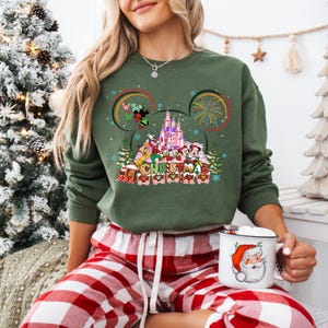 May include: Green Christmas sweatshirt featuring a Disney castle and characters, including Mickey and Minnie Mouse. The design incorporates fireworks, snowflakes, and the word "CHRISTMAS" in a festive font. The sweatshirt is paired with red and white plaid pajama pants.
