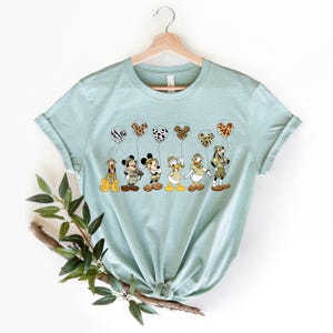 Pode incluir: Uma camiseta azul claro com um gr&aacute;fico de Mickey Mouse, Donald Duck, Pateta, Pluto e alguns outros personagens da Disney segurando bal&otilde;es com estampa de animais. Os personagens est&atilde;o usando roupas de saf&aacute;ri.