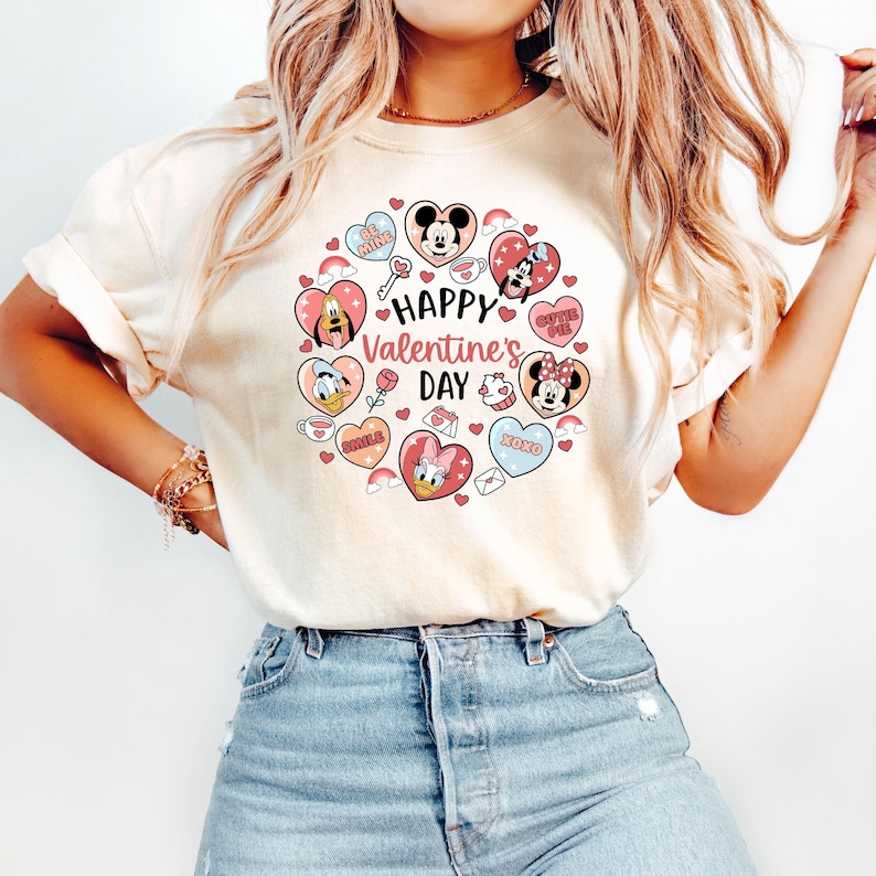 K&ouml;nnte beinhalten: Cremefarbenes T-Shirt mit einem Valentinstagsdesign. Das Design umfasst herzf&ouml;rmige Illustrationen von Disney-Charakteren wie Mickey und Minnie Mouse, Donald Duck und Daisy Duck, mit dem Text "Happy Valentine's Day".