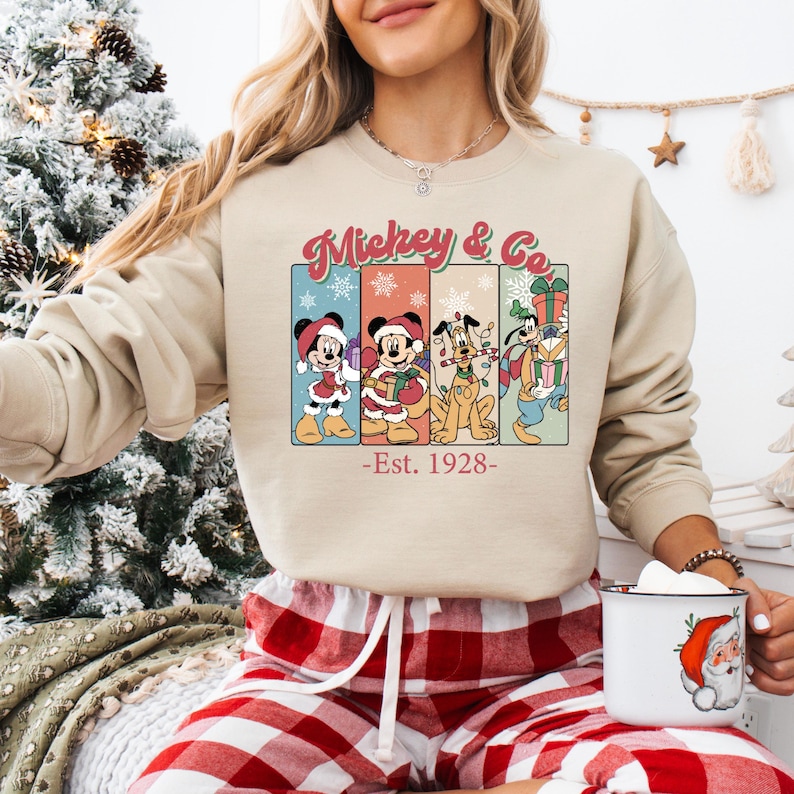 May include: Beige sweatshirt with a graphic featuring Mickey Mouse and friends in holiday attire. The text "Mickey & Co" is above the characters, with "-Est. 1928-" below. The person is wearing red and white plaid pajama pants.
