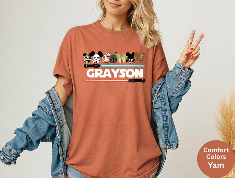 May include: A rust-colored t-shirt featuring a graphic design with cartoon character heads, lightsabers, and the name "GRAYSON" in white block letters. The shirt has short sleeves and a crew neck.