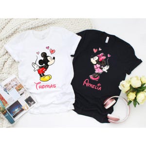 May include: Two t-shirts, one white and one black, each featuring a cartoon character design. The white shirt has a Mickey Mouse graphic with the name "Thomas" below. The black shirt features a Minnie Mouse graphic with the name "Amelia".
