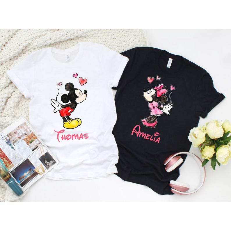 May include: Two t-shirts, one white and one black, each featuring a cartoon character design. The white shirt has a Mickey Mouse graphic with the name "Thomas" below. The black shirt features a Minnie Mouse graphic with the name "Amelia".