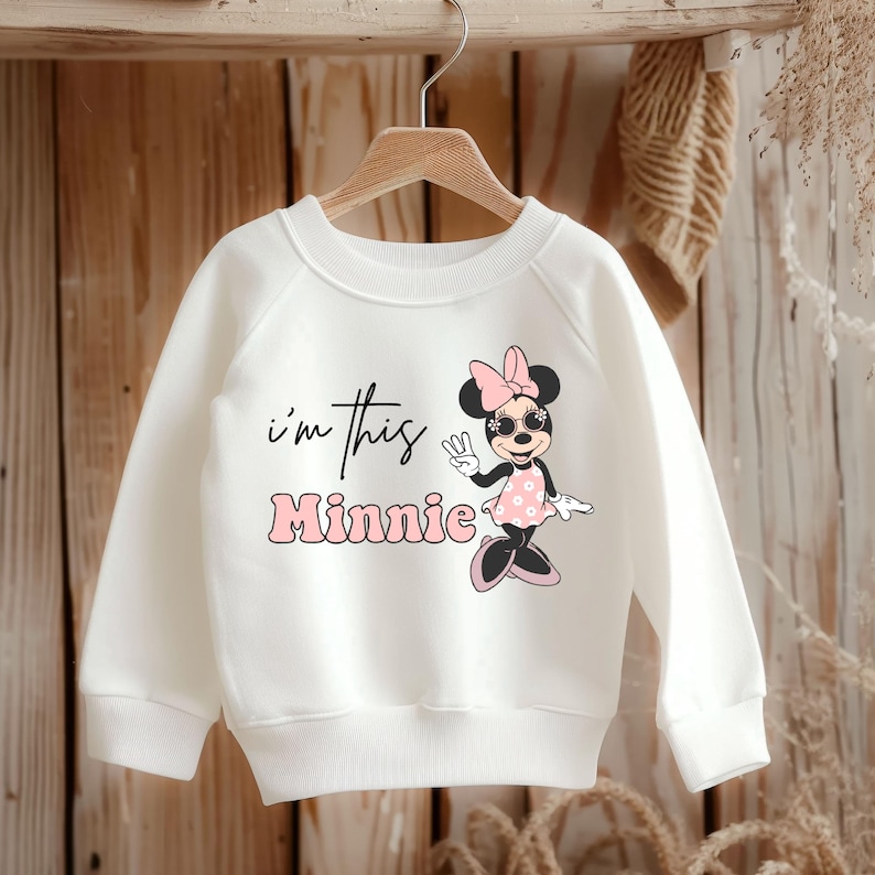 Puede incluir: Sudadera blanca con un gr&aacute;fico rosa de Minnie Mouse y el texto "i'm this Minnie" en negro.