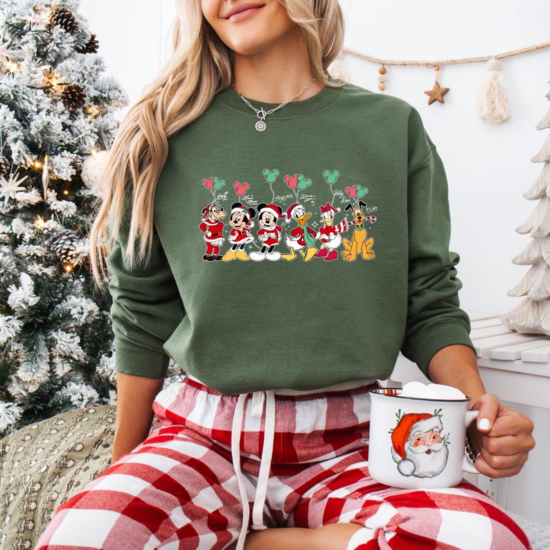 May include: Green sweatshirt featuring a festive Disney character design. The graphic includes Mickey Mouse, Minnie Mouse, Donald Duck, and Pluto in holiday attire, holding balloons. The person is also wearing red and white plaid pajama pants and holding a Santa mug.