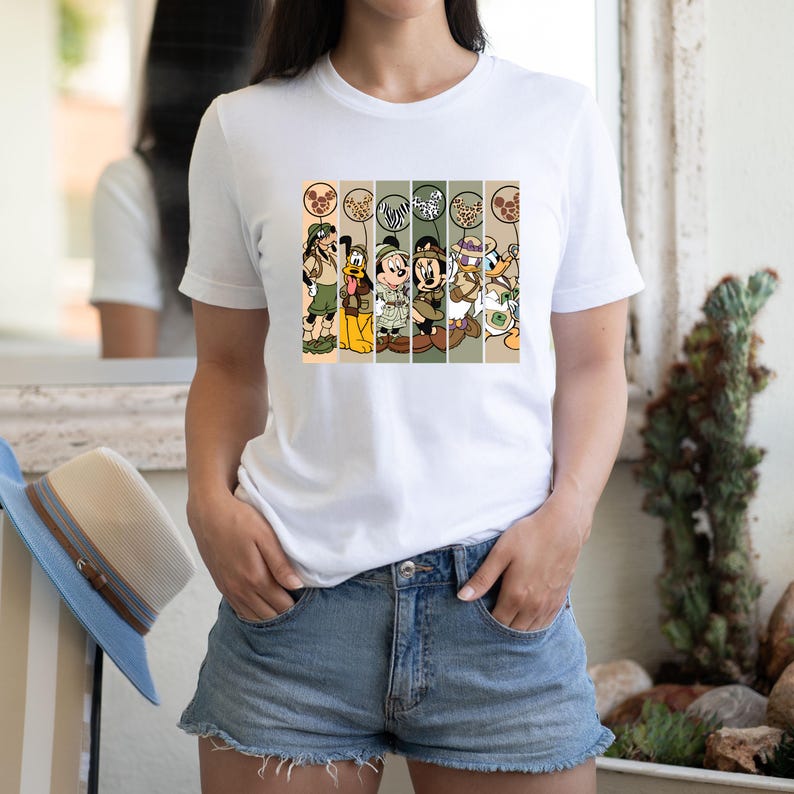 May include: White t-shirt with a colorful graphic print of Disney characters dressed in safari outfits. The characters include Mickey Mouse, Minnie Mouse, Donald Duck, Goofy, and Pluto. The graphic is set against a background of stripes and animal print.