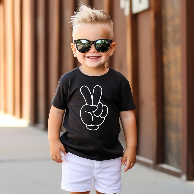 K&ouml;nnte beinhalten: Ein schwarzes T-Shirt mit einem wei&szlig;en Peace-Zeichen-Motiv. Das T-Shirt wird mit wei&szlig;en Shorts kombiniert. Die Person tr&auml;gt eine schwarze Sonnenbrille und l&auml;chelt. Das Outfit ist l&auml;ssig und f&uuml;r Kinder geeignet.