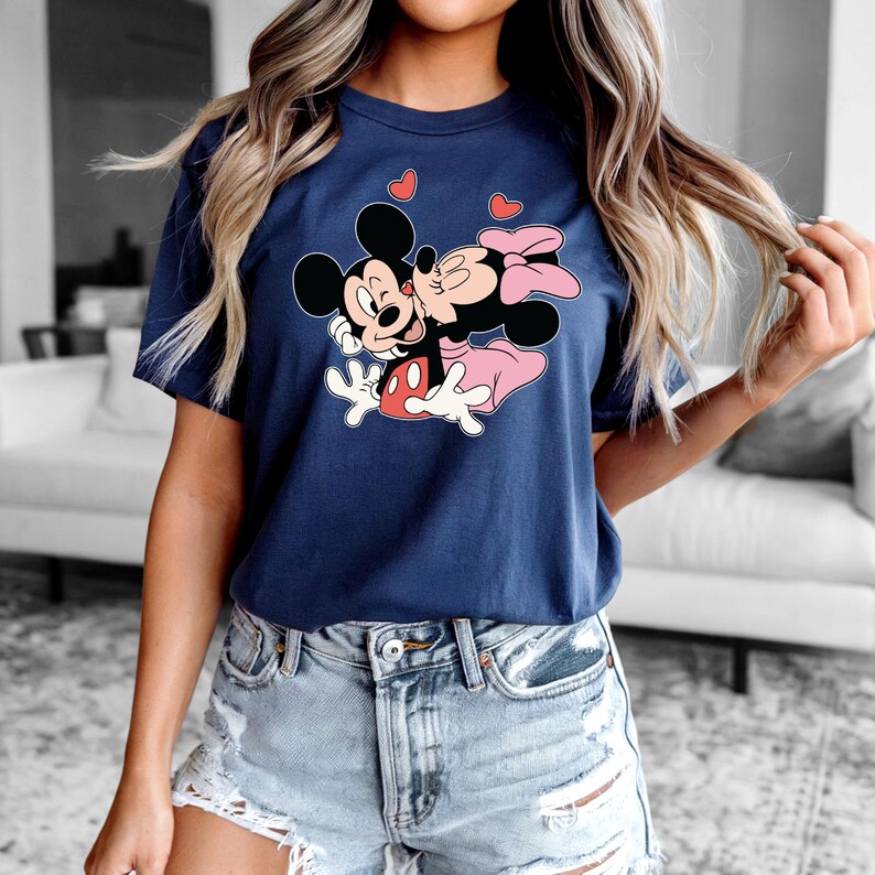 May include: Navy blue t-shirt featuring a graphic of Mickey and Minnie Mouse embracing, with red hearts above them. The cartoon characters are in black, white, red, and pink. The shirt is a short-sleeved crew neck.