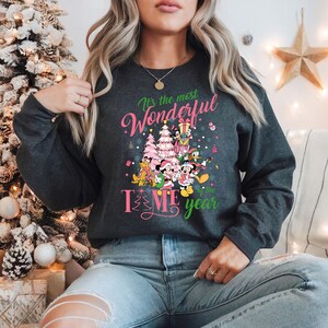 May include: A gray sweatshirt with a pink and green Christmas design featuring Mickey Mouse and friends. The text on the sweatshirt reads "It's the most Wonderful Time of the Year".