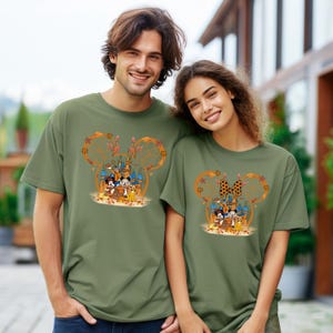 Pode incluir: Duas camisetas verde-oliva com um design em forma de Mickey Mouse com um castelo, personagens e folhas de outono. O design inclui Mickey e Minnie Mouse, Pato Donald e outros personagens da Disney. As camisetas s&atilde;o de manga curta.