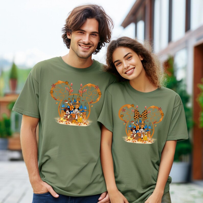 Pode incluir: Duas camisetas verde-oliva com um design em forma de Mickey Mouse com um castelo, personagens e folhas de outono. O design inclui Mickey e Minnie Mouse, Pato Donald e outros personagens da Disney. As camisetas s&atilde;o de manga curta.
