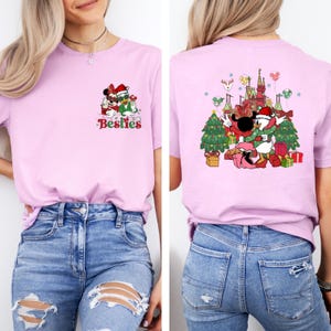 May include: A light pink t-shirt featuring a Christmas-themed graphic of Disney characters. The front of the shirt says "Besties" in red and green. The back of the shirt has a larger design with a castle, Christmas trees, and presents.
