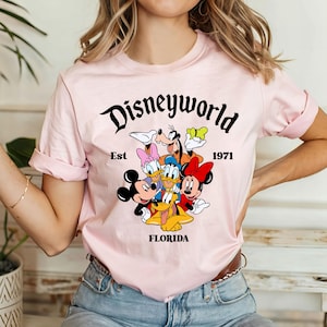 Pu&ograve; includere: Maglietta rosa con una grafica nera di Topolino, Minnie Mouse, Paperino, Pippo e Pluto. La grafica include il testo "Disneyworld Est 1971 Florida".