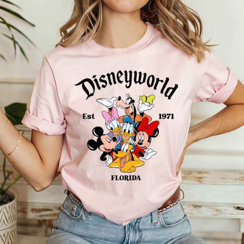 Pu&ograve; includere: Maglietta rosa con una grafica nera di Topolino, Minnie Mouse, Paperino, Pippo e Pluto. La grafica include il testo "Disneyworld Est 1971 Florida".