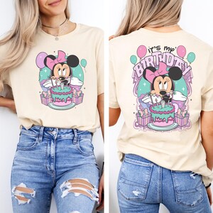 May include: A beige t-shirt with a graphic of Minnie Mouse holding a birthday cake with candles. The back of the shirt says "it's my birthday" with a graphic of Minnie Mouse holding a birthday cake with candles.