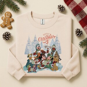 May include: Cream-colored sweatshirt featuring a graphic of Disney characters singing Christmas carols in a snowy scene. The phrase "Merry Christmas" is written above the characters. The sweatshirt has long sleeves and a crew neck.