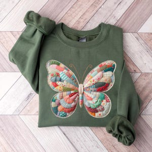 May include: A green sweatshirt featuring a large, colorful butterfly applique. The butterfly design is made of various fabric patches in shades of red, blue, green, and yellow. The sleeves are cuffed, and the sweatshirt is folded on a wooden surface.